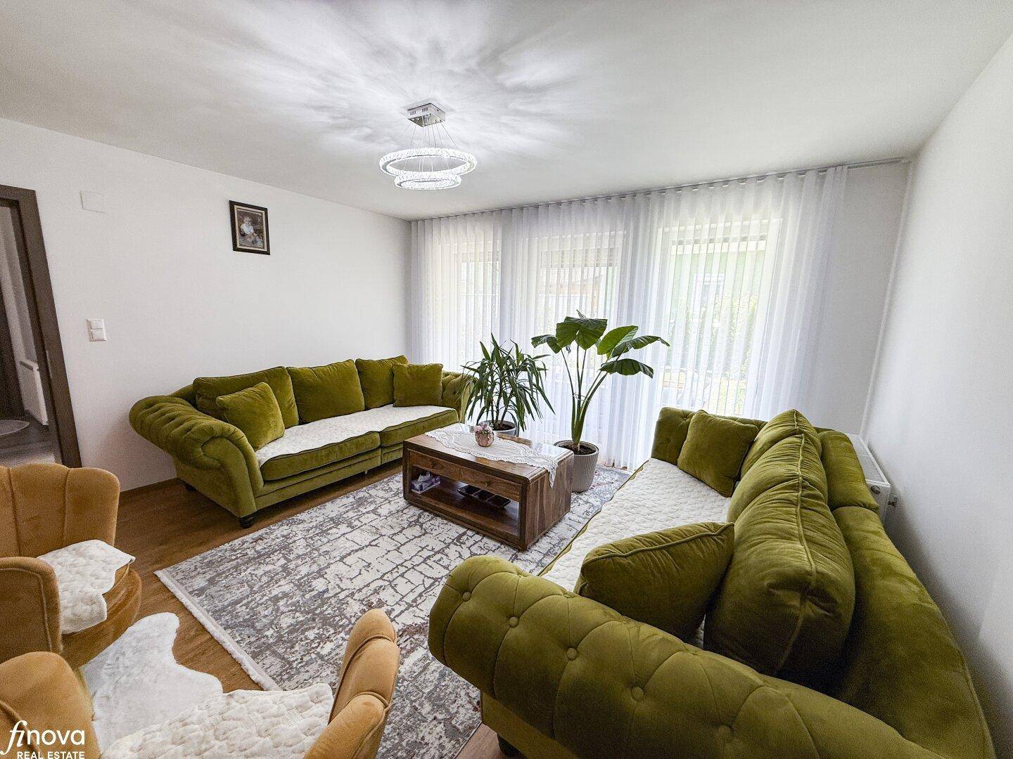 Lichtdurchflutetes Wohnzimmer mit großen Fenstern, grünen Sofas und Pflanzen.