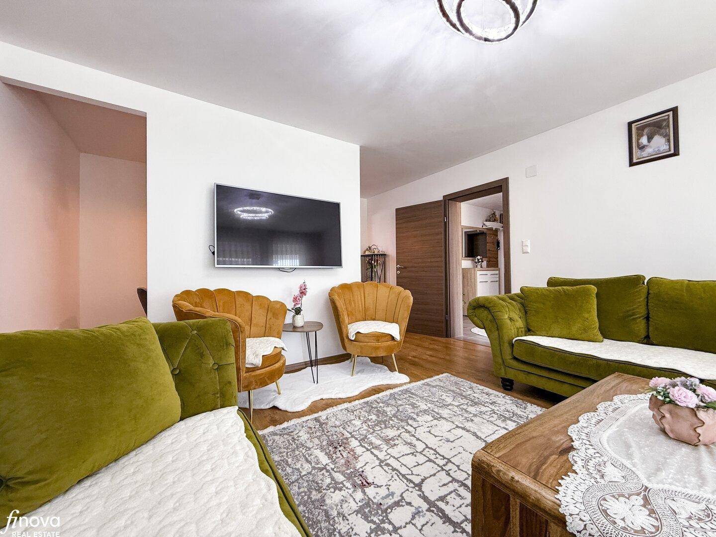 Gemütliches Wohnzimmer mit Wandfernseher, bequemen Sitzgelegenheiten und Holzboden.