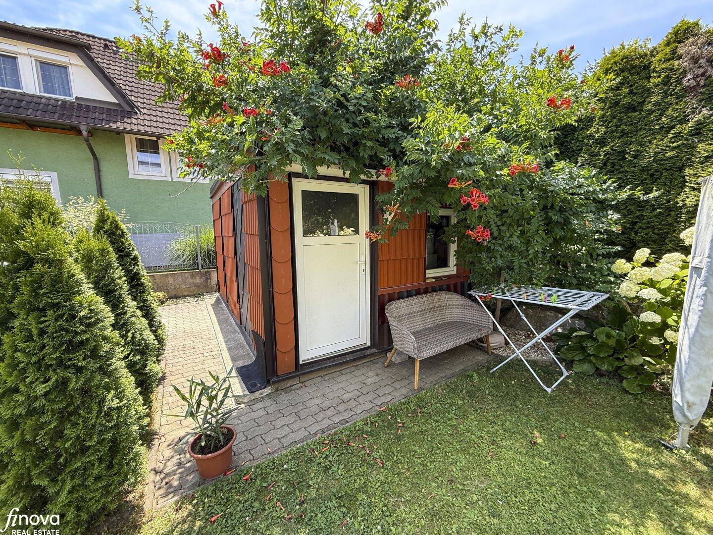 Kleiner Gartenbereich mit einem Holzschuppen, Sitzgelegenheit und Wäscheständer umgeben von Pflanzen.