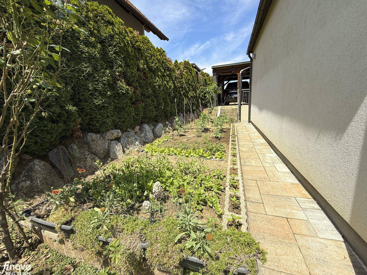 Seitlicher Gartenbereich mit Hochbeeten, einem gepflasterten Weg und einer Hecke im Hintergrund.
