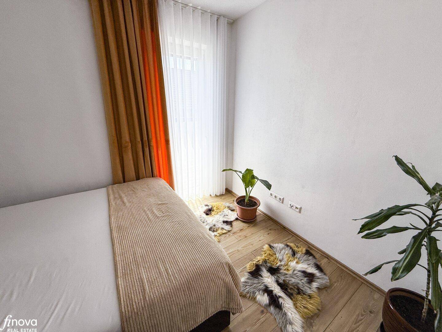 Schlafzimmer mit Holzboden, Pflanzen und einem Fenster mit hellen Vorhängen.