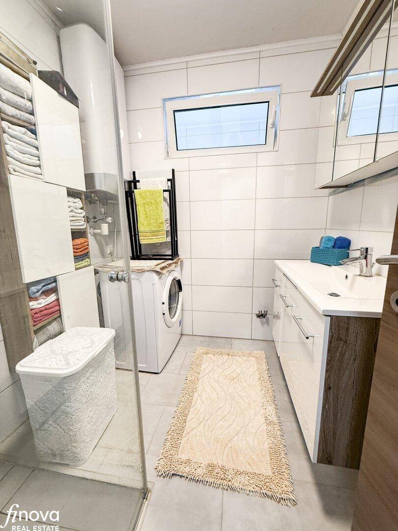 Helles Badezimmer mit weißen Fliesen, Duschkabine, Waschbecken und Waschmaschine.