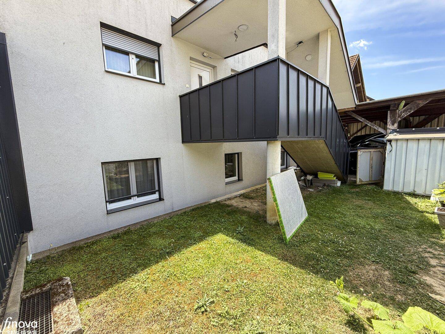 Außenansicht des Gebäudes mit Rasenfläche, einem Balkon mit schwarzem Geländer und Rollläden.