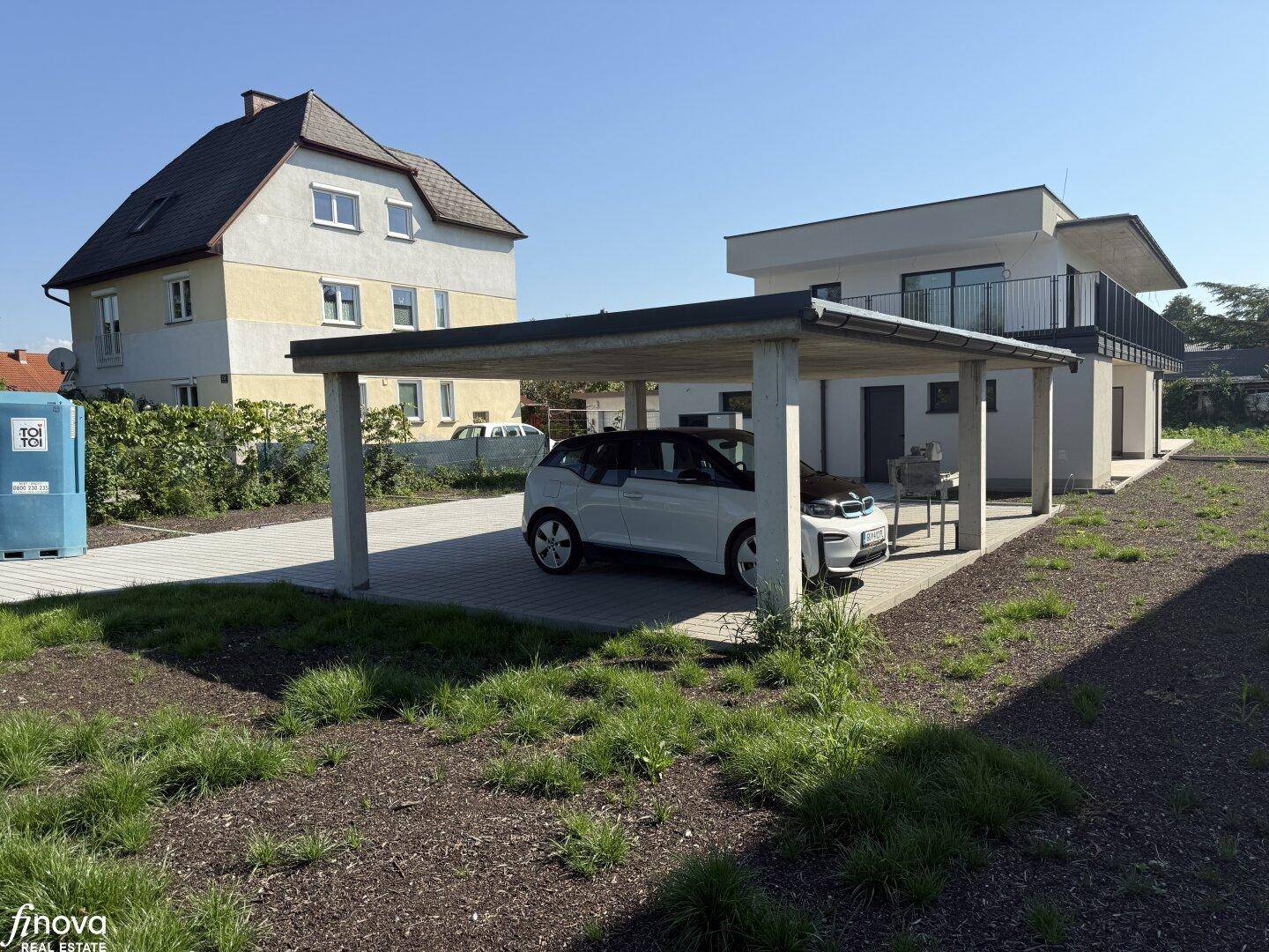 Neubau-Doppelhaushälfte mit Carport und einem Elektrofahrzeug auf dem Grundstück, umgeben von Grünflächen.