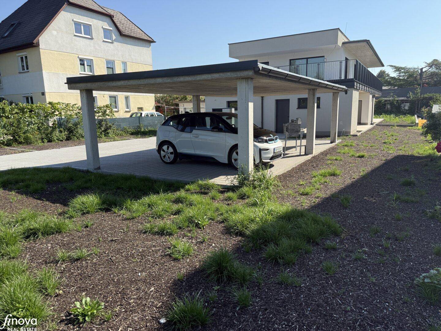 Carport mit geparktem Auto neben dem modernen Gebäude und einem unbebauten Gartenbereich.