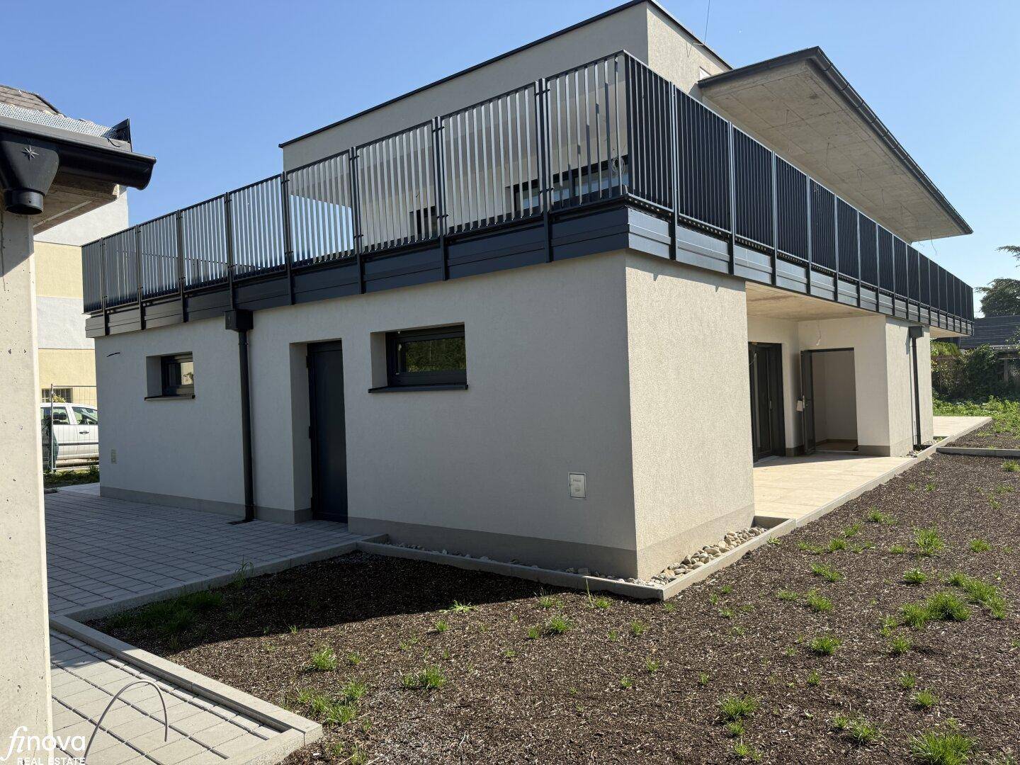 Seitliche Ansicht des modernen Gebäudes mit einer gepflasterten Terrasse und dem darüberliegenden Balkon.