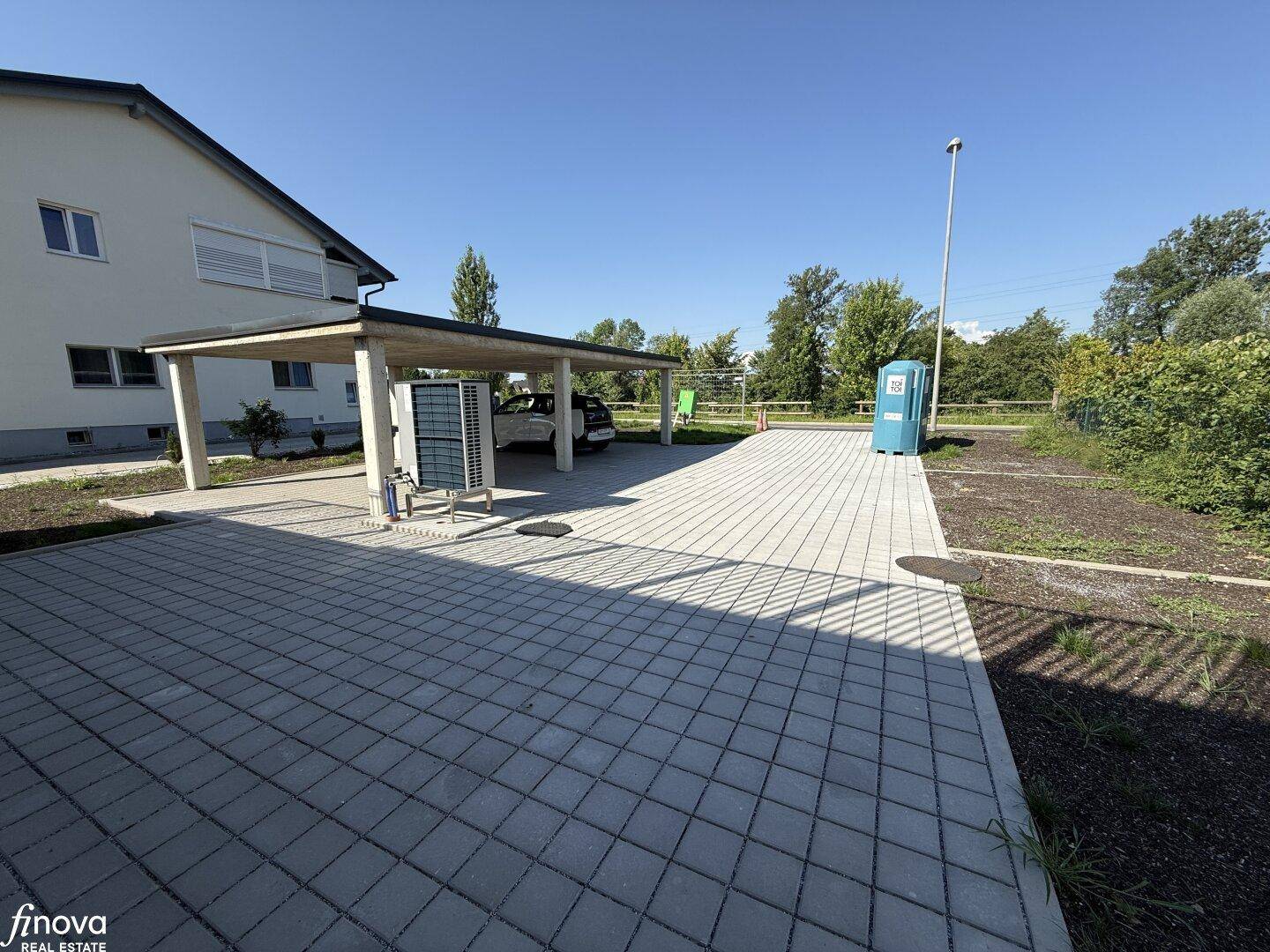 Weitläufiger Außenbereich mit Carport, gepflasterter Zufahrt und unbebautem Gartenland neben dem Gebäude.