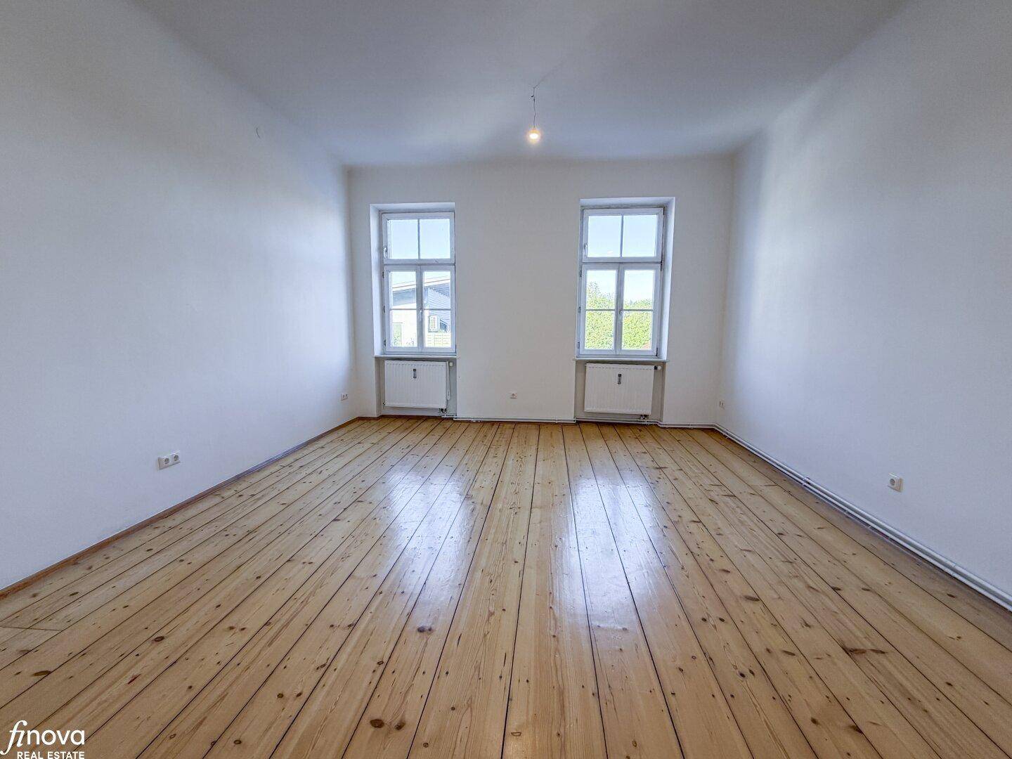 Lichtdurchfluteter Raum mit zwei großen Fenstern und glänzendem Holzdielenboden, sehr einladend.
