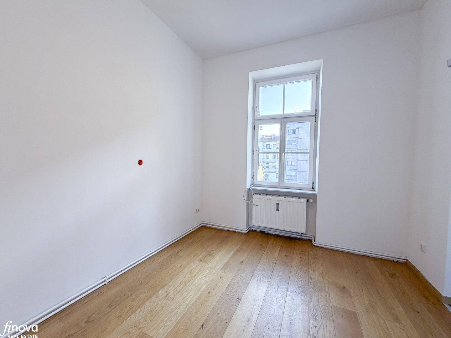 Helles Zimmer mit Holzboden und Fensterblick auf die Stadt, ausgestattet mit Heizkörper.