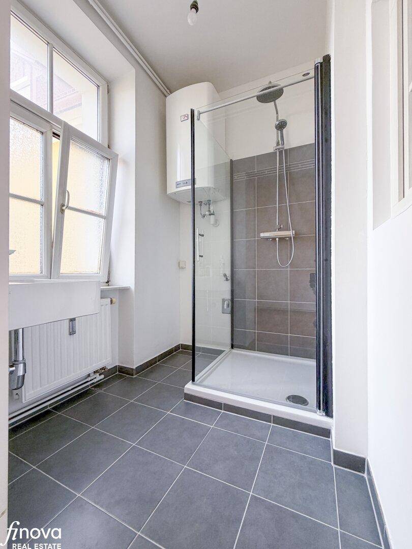 Modernes Duschbad mit grauen Fliesen, Fenster und Warmwasserboiler, sehr funktional.