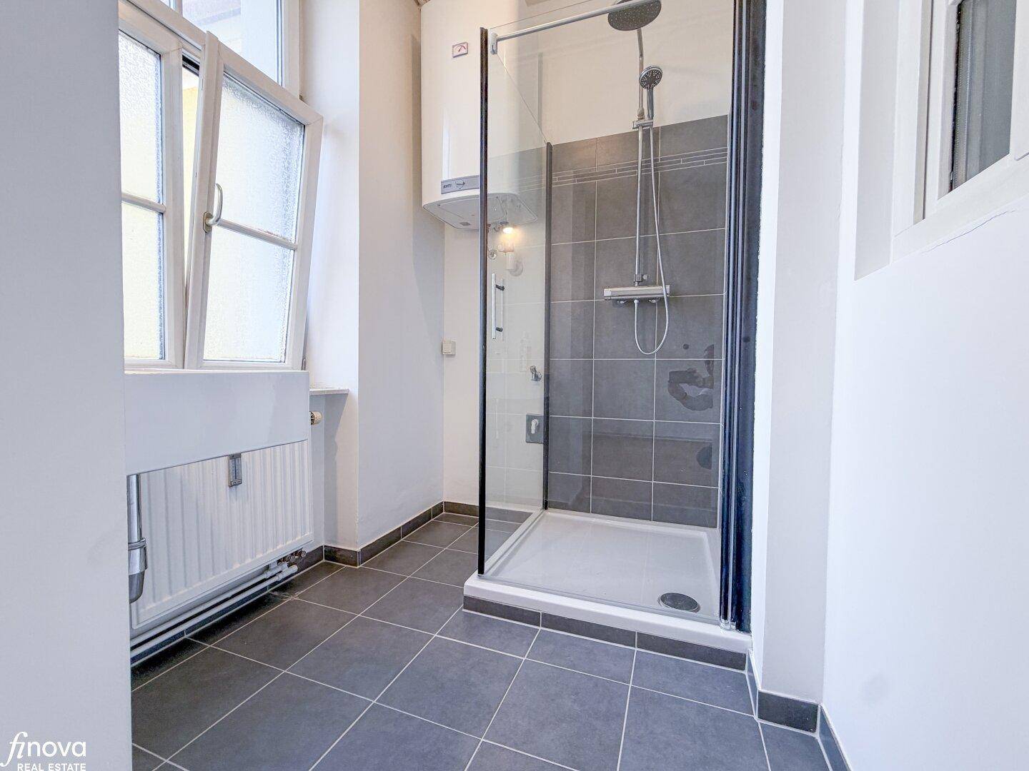 Funktionales Duschbad mit grauen Fliesen, Fenster und Warmwasserboiler, bietet ausreichend Platz.