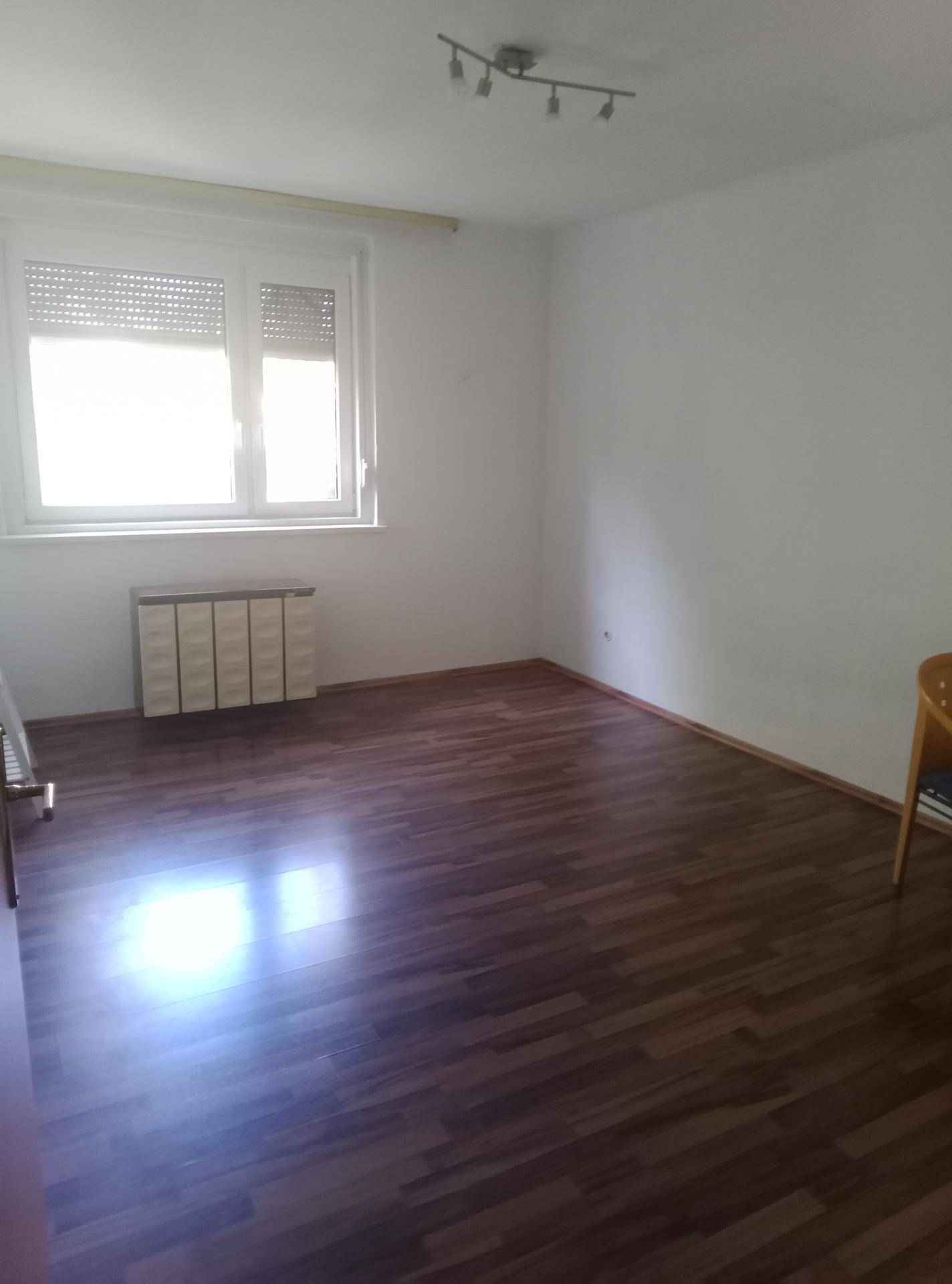 Leeres Zimmer mit Laminatboden, weißen Wänden und einem Fenster mit Außenjalousie.
