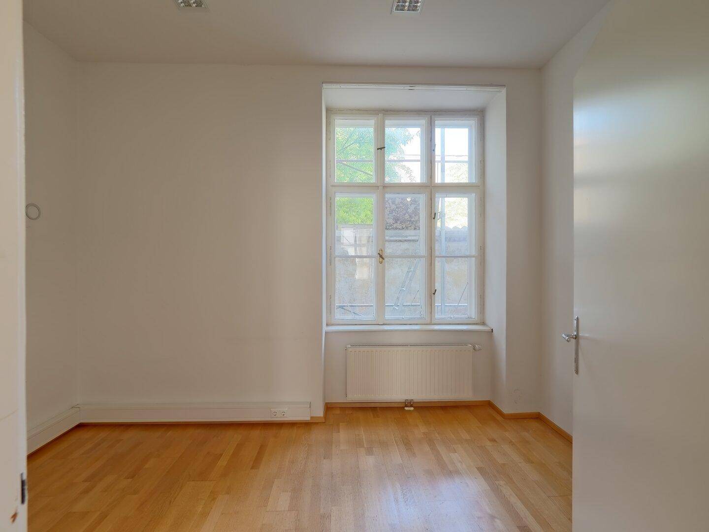 Leerer Büroraum mit einem großen Fenster, Heizkörper und hellem Holzparkettboden, bereit zur Einrichtung.