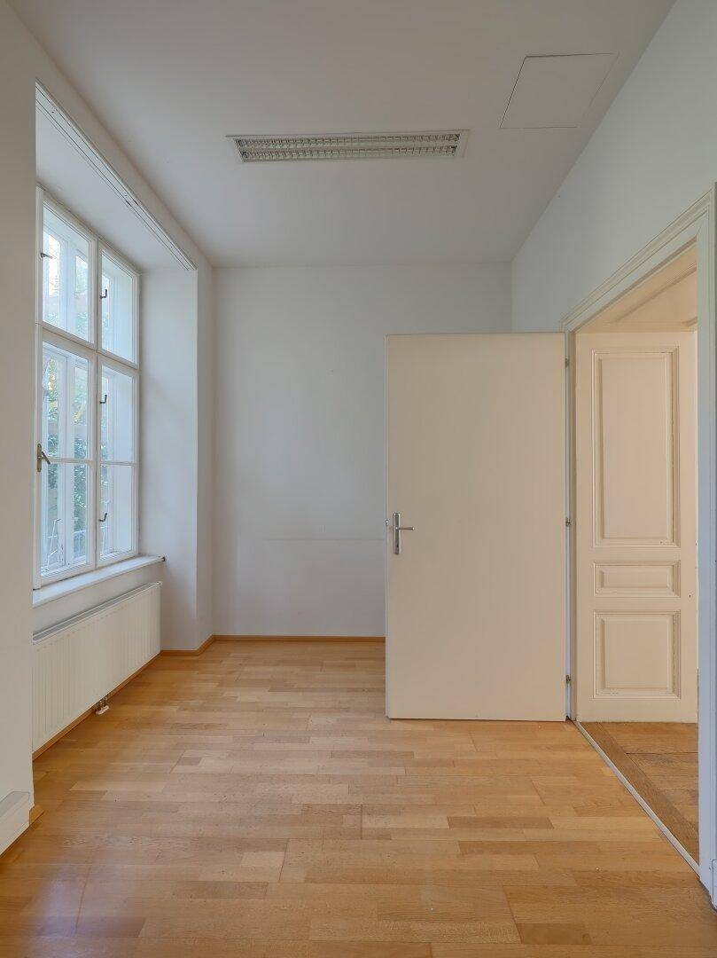 Heller Büroraum mit Holzboden, großen Fenstern und einer offenen Tür zu einem angrenzenden Bereich.