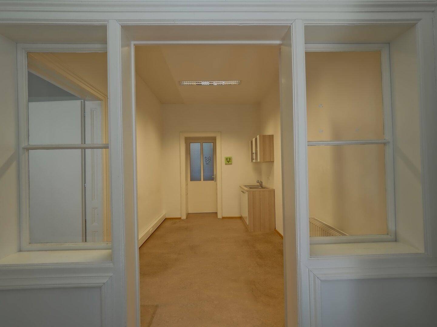 Blick durch eine Türöffnung in einen Raum mit Teppichboden und einer kleinen Küchenzeile.