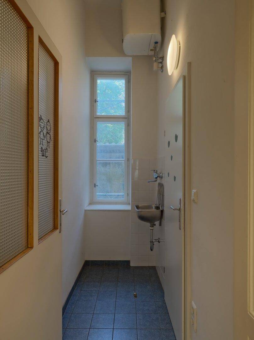 Schmaler Waschraum mit kleinem Waschbecken, Fenster und dunklen Fliesen auf dem Boden.