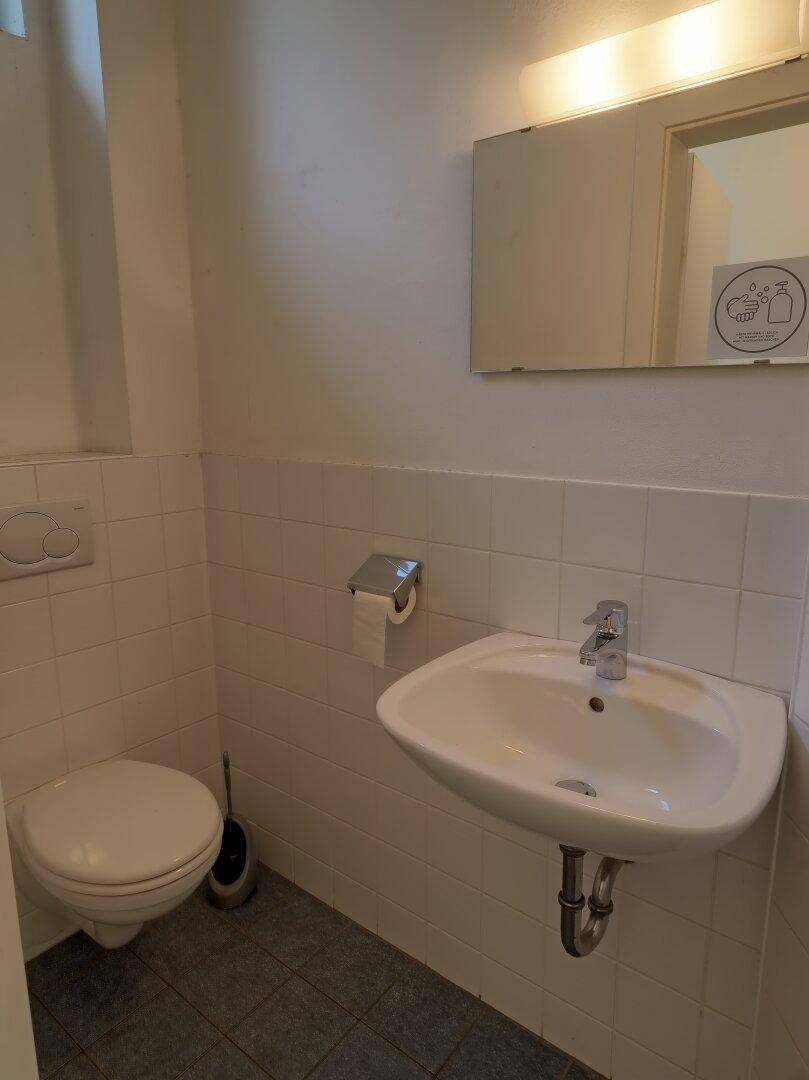 Toilettenraum mit wandhängendem WC, Waschbecken, Spiegel und weißen Wandfliesen.