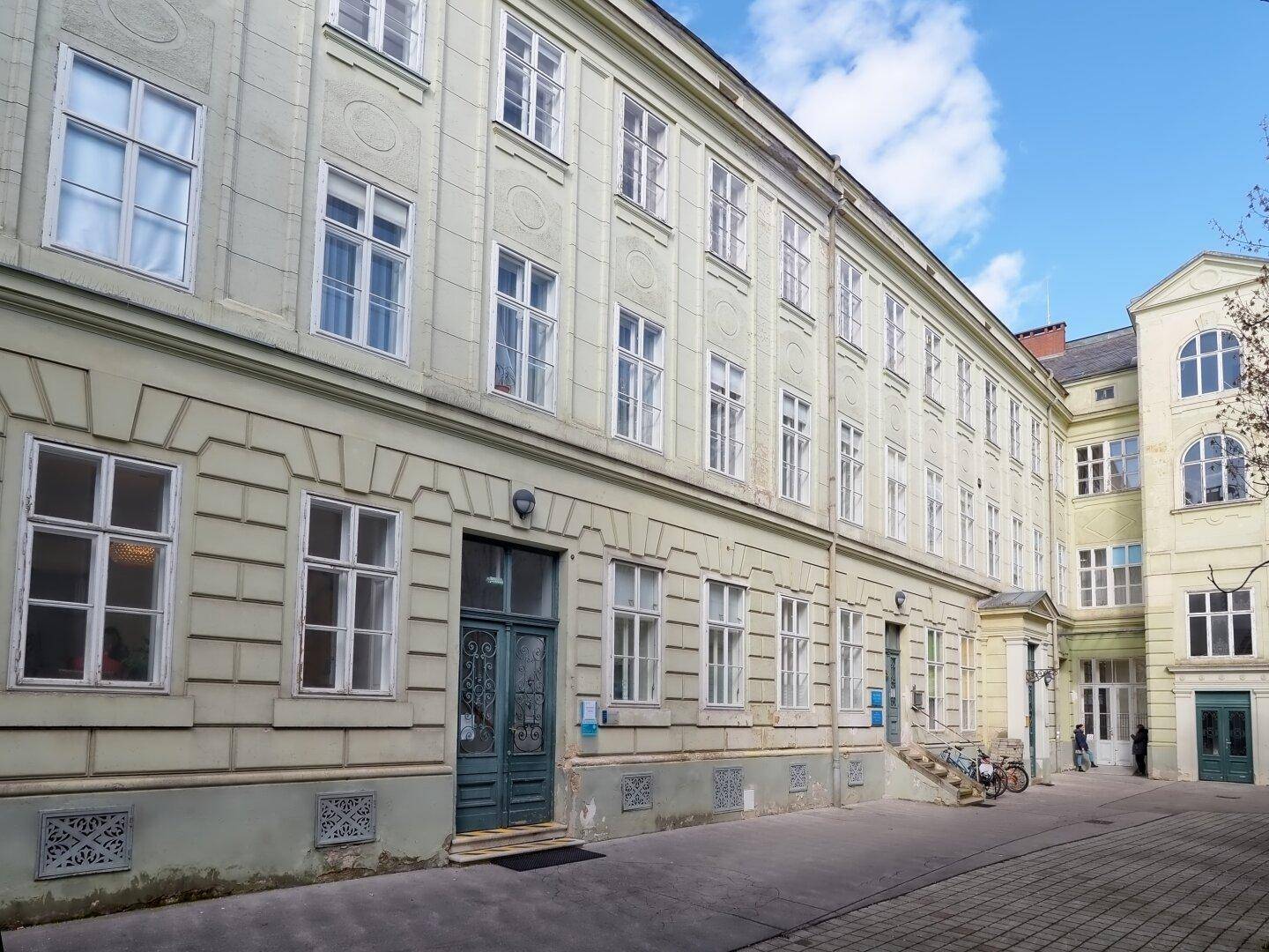 Historisches Gebäude mit vielen Fenstern, einer grünen Fassade und einem gepflasterten Eingangsbereich.