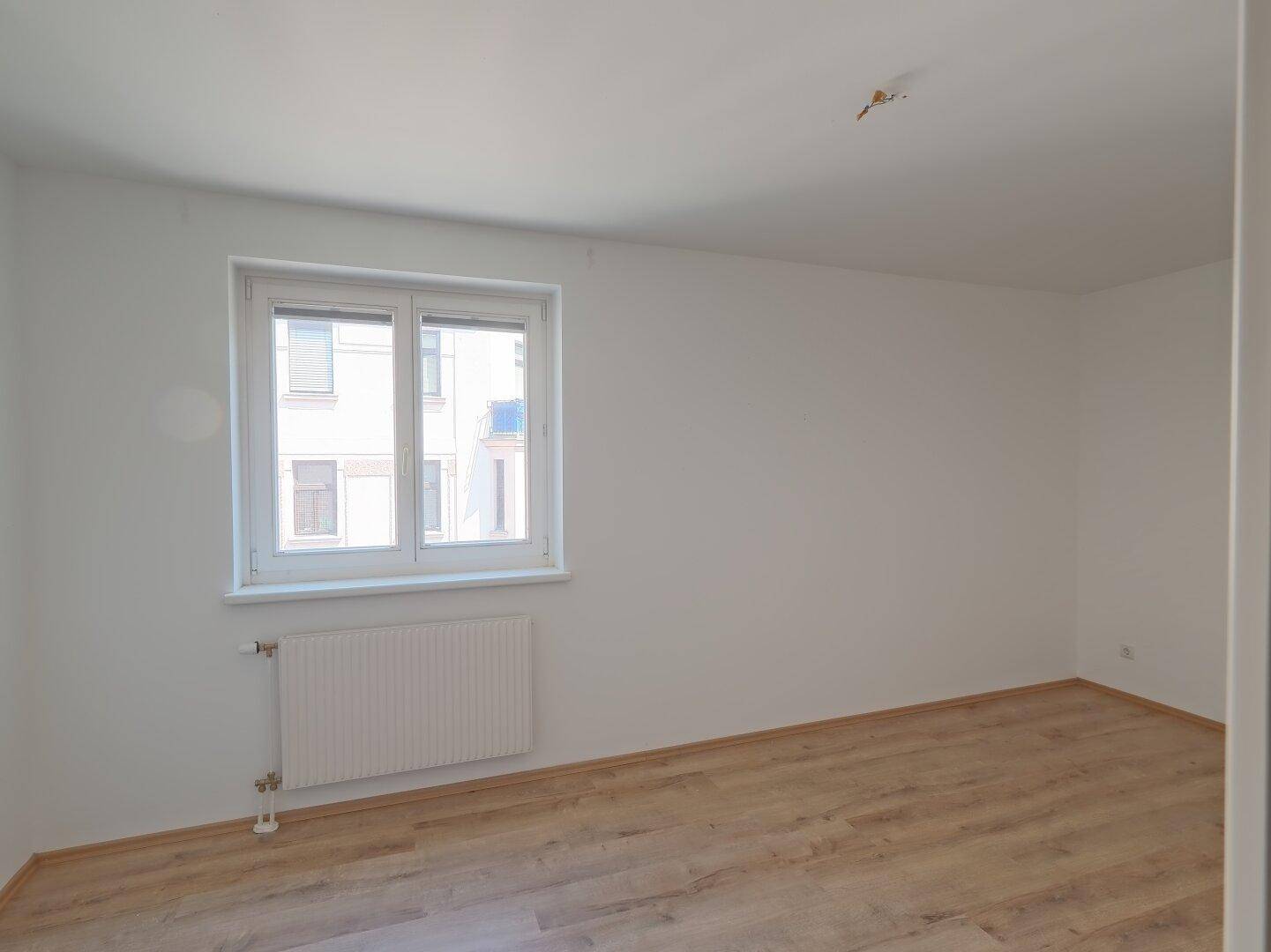 Leeres Schlafzimmer mit Laminatboden, einem Fenster und Blick auf die umliegenden Gebäude.