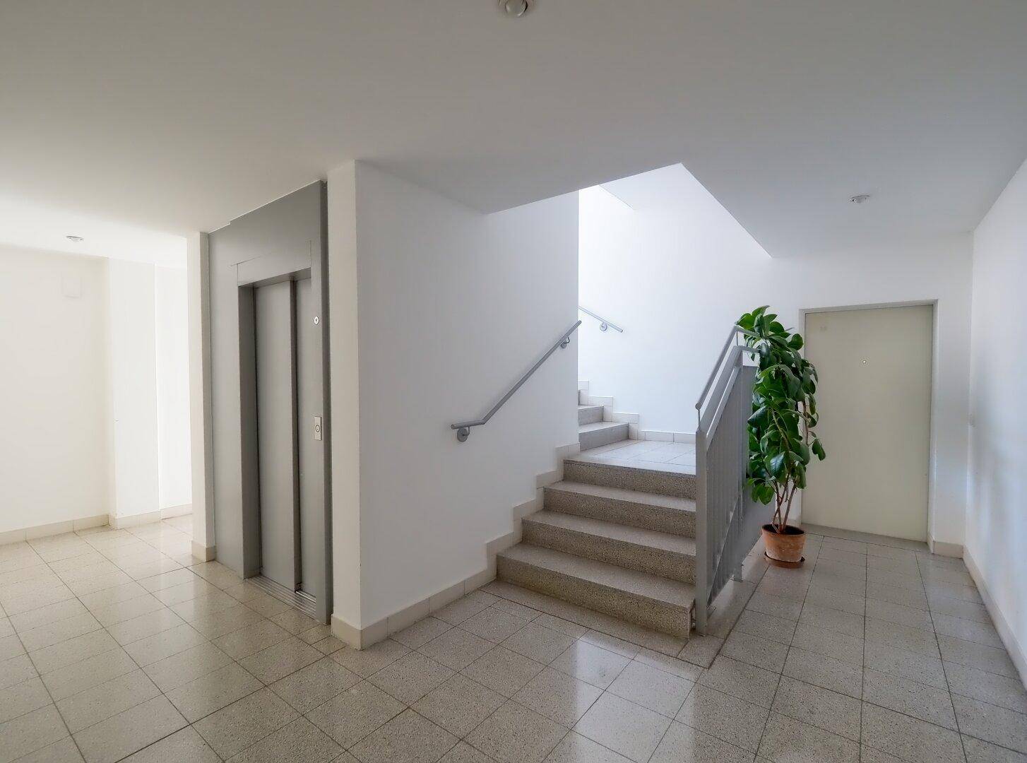 Heller Eingangsbereich mit Treppenhaus, Aufzug und gefliestem Boden in einem Mehrfamilienhaus.