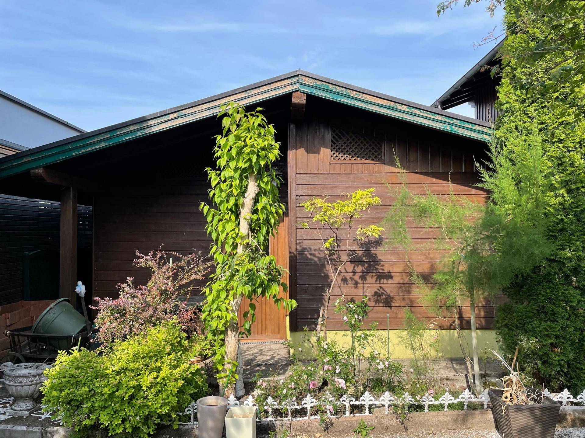 Kleines Holzhaus im Garten, umgeben von Sträuchern und Bäumen unter blauem Himmel.