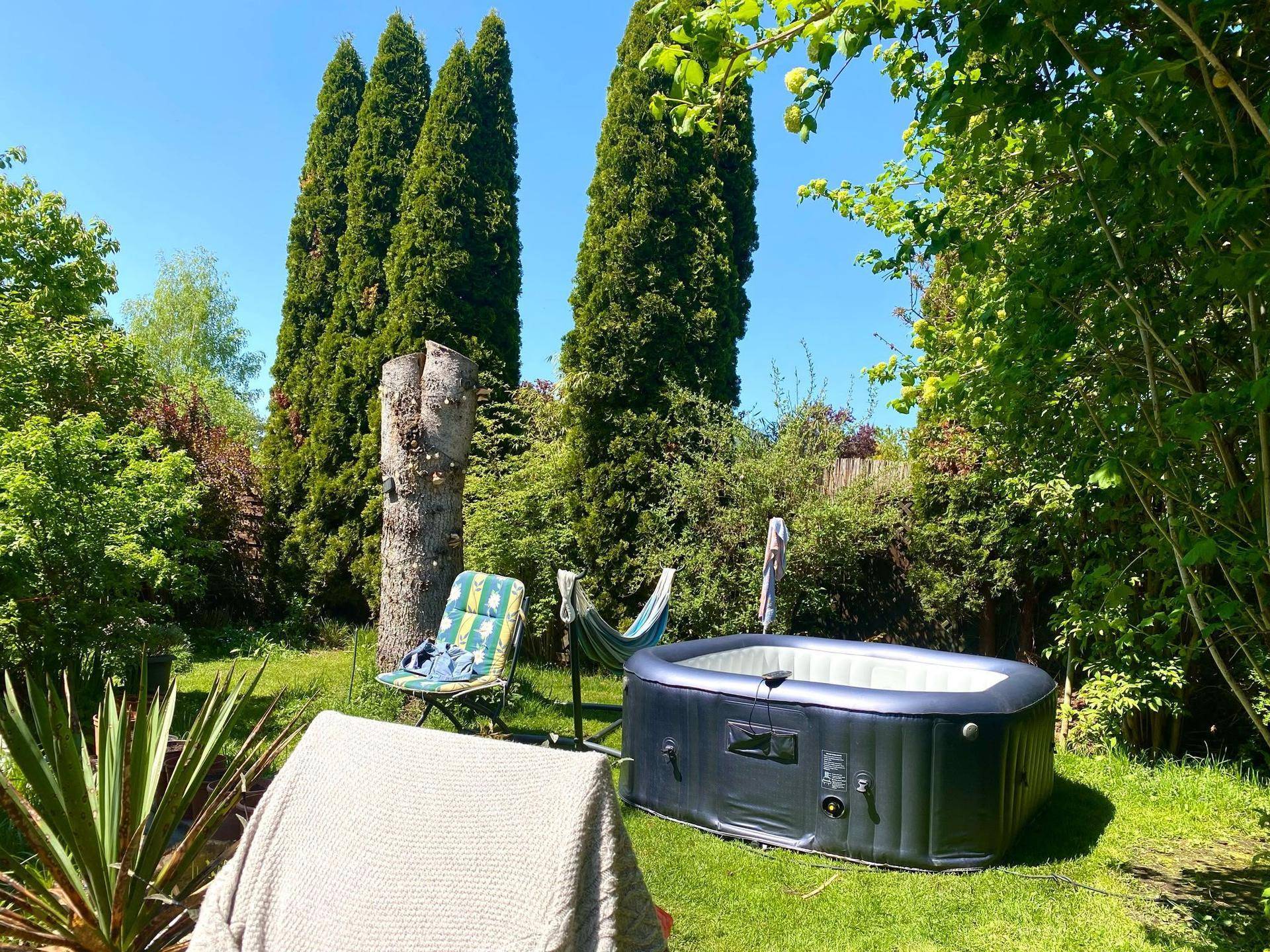 Sonniger Garten mit aufblasbarem Whirlpool, Liegestuhl und hohen Zypressen.