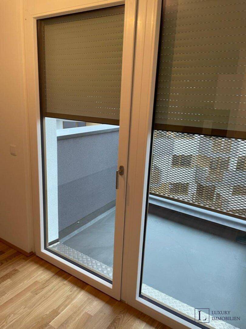 Fenster mit heruntergelassenem Rollladen und Blick auf den Außenbereich des Balkons.