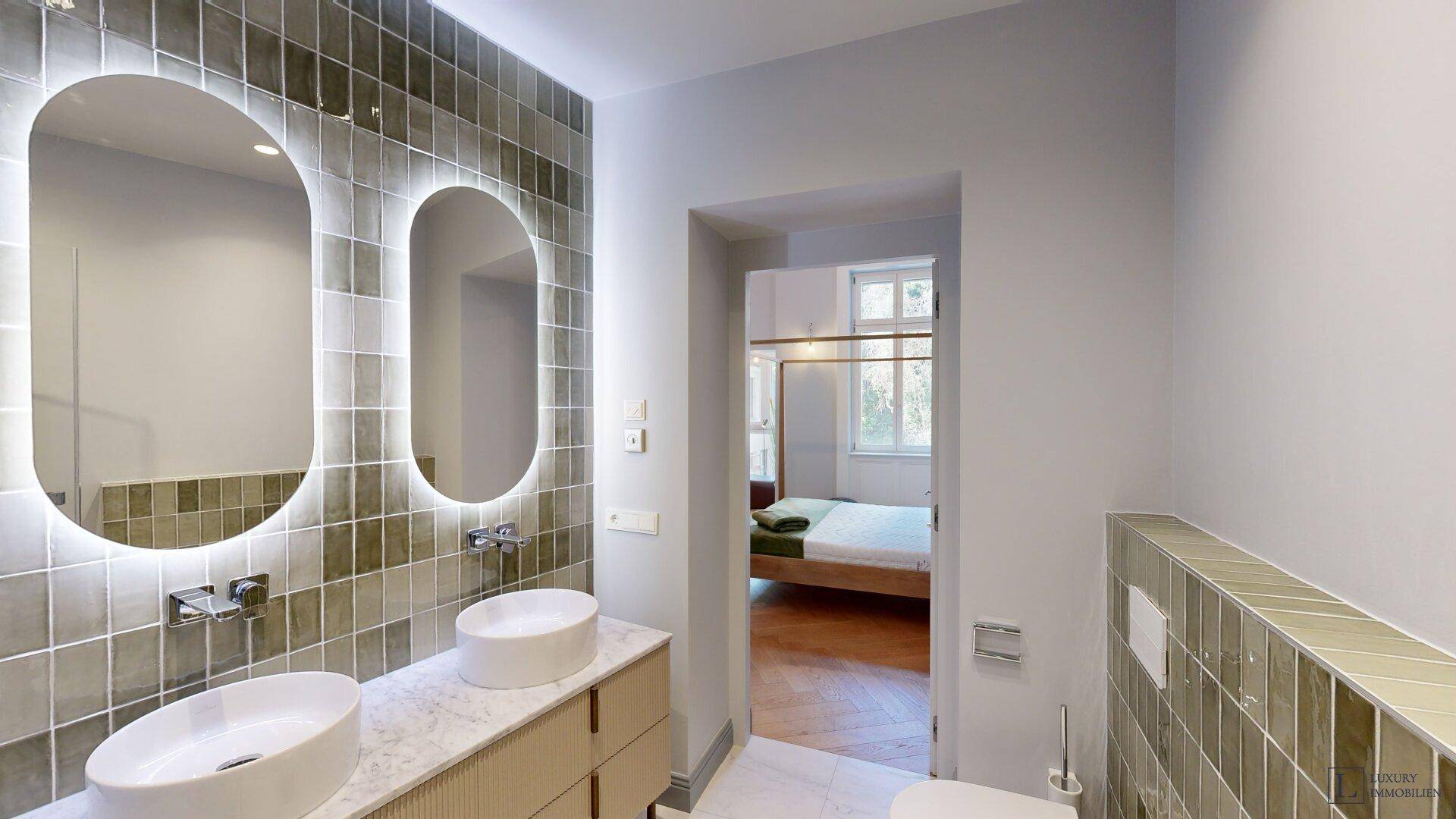 Modernes Badezimmer mit Doppelwaschbecken, beleuchteten Spiegeln und grünen Fliesen.