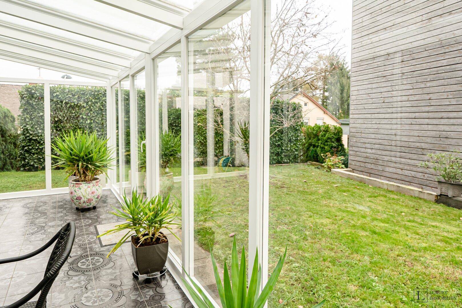 Grüner Gartenbereich mit Rasenfläche und Bäumen, gesehen durch die Glasfront des Wintergartens.
