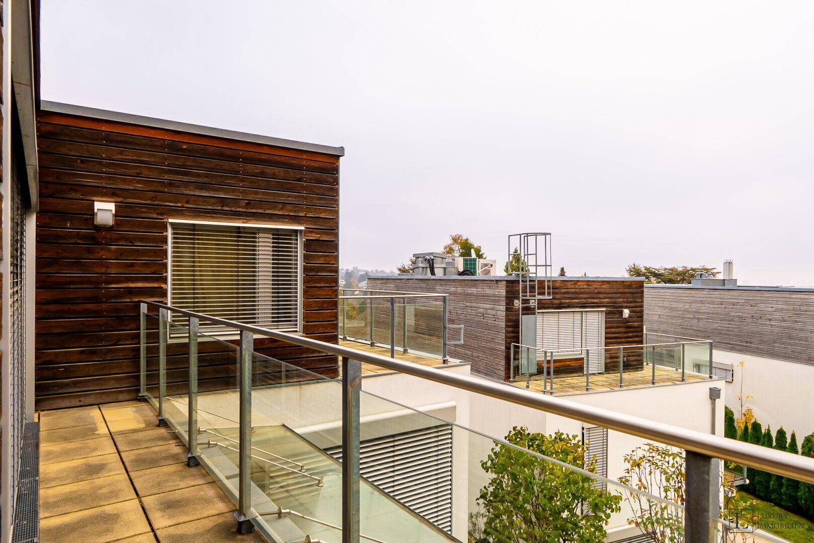 Langer Balkon mit Glasgeländer und weitem Blick über die umliegenden Gebäude und Natur.