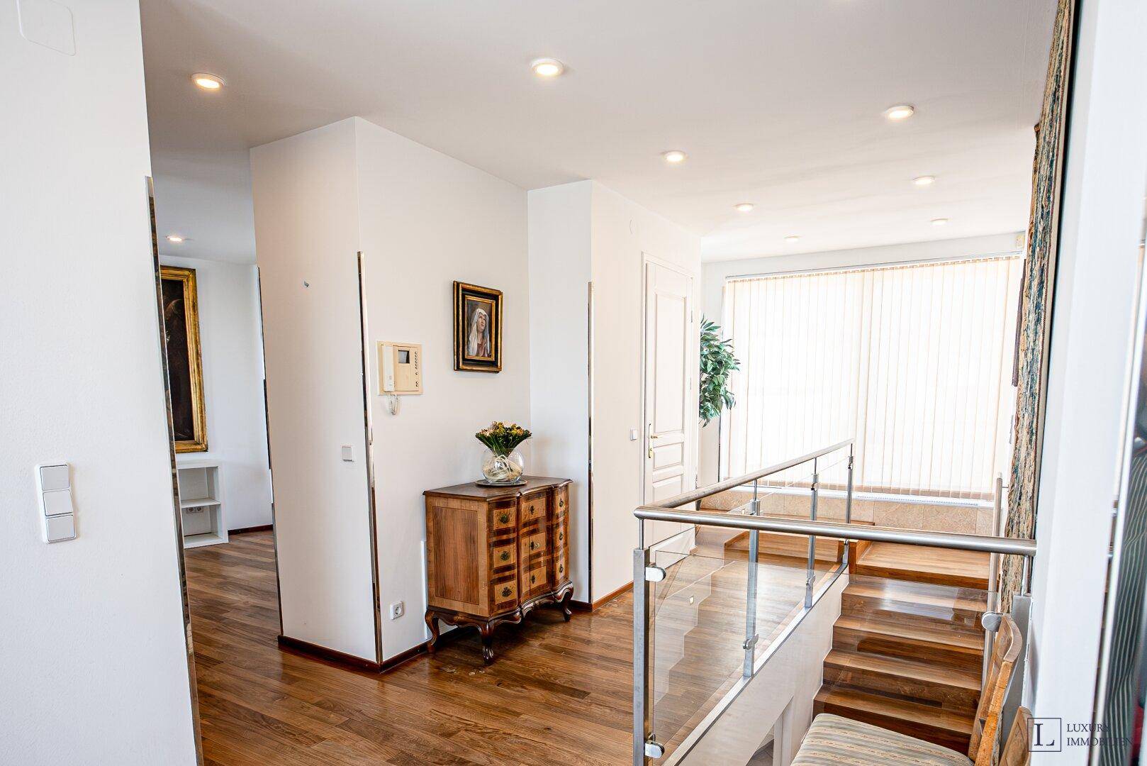 Heller Eingangsbereich mit Holzparkettboden, Einbaustrahlern und Treppe mit Glasgeländer.
