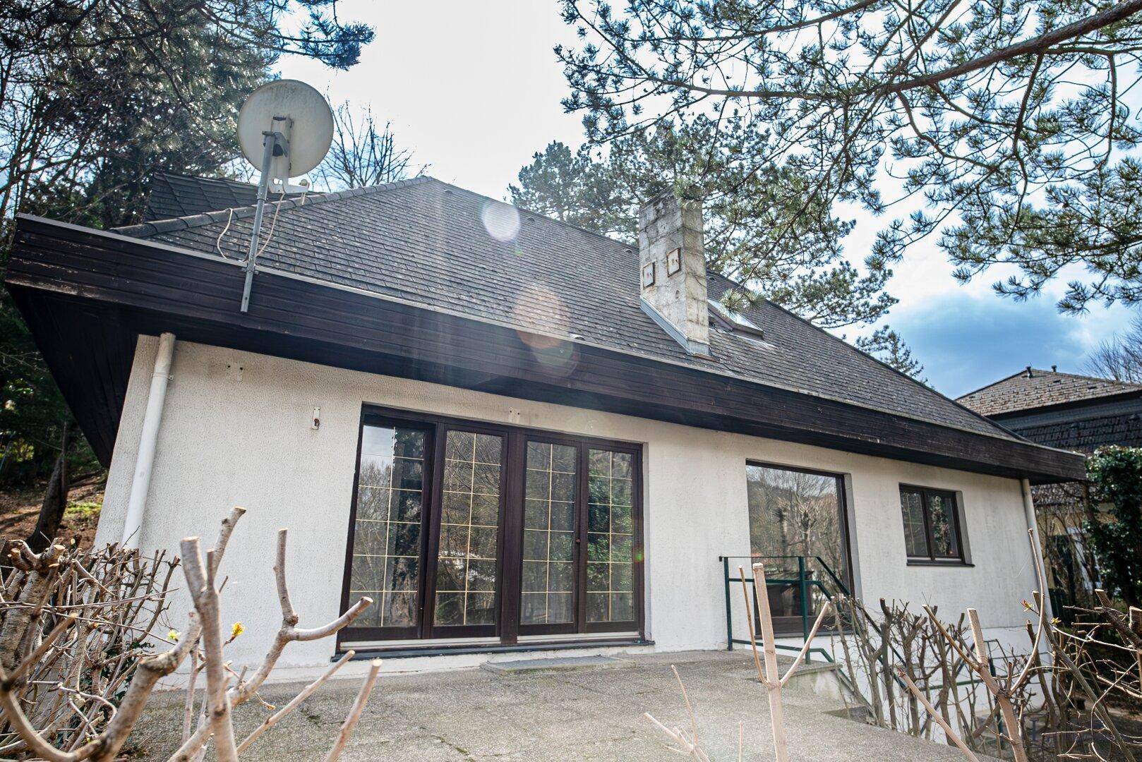 Rückseite des Hauses mit einer gepflasterten Terrasse, großen Fenstern und einem Kamin.