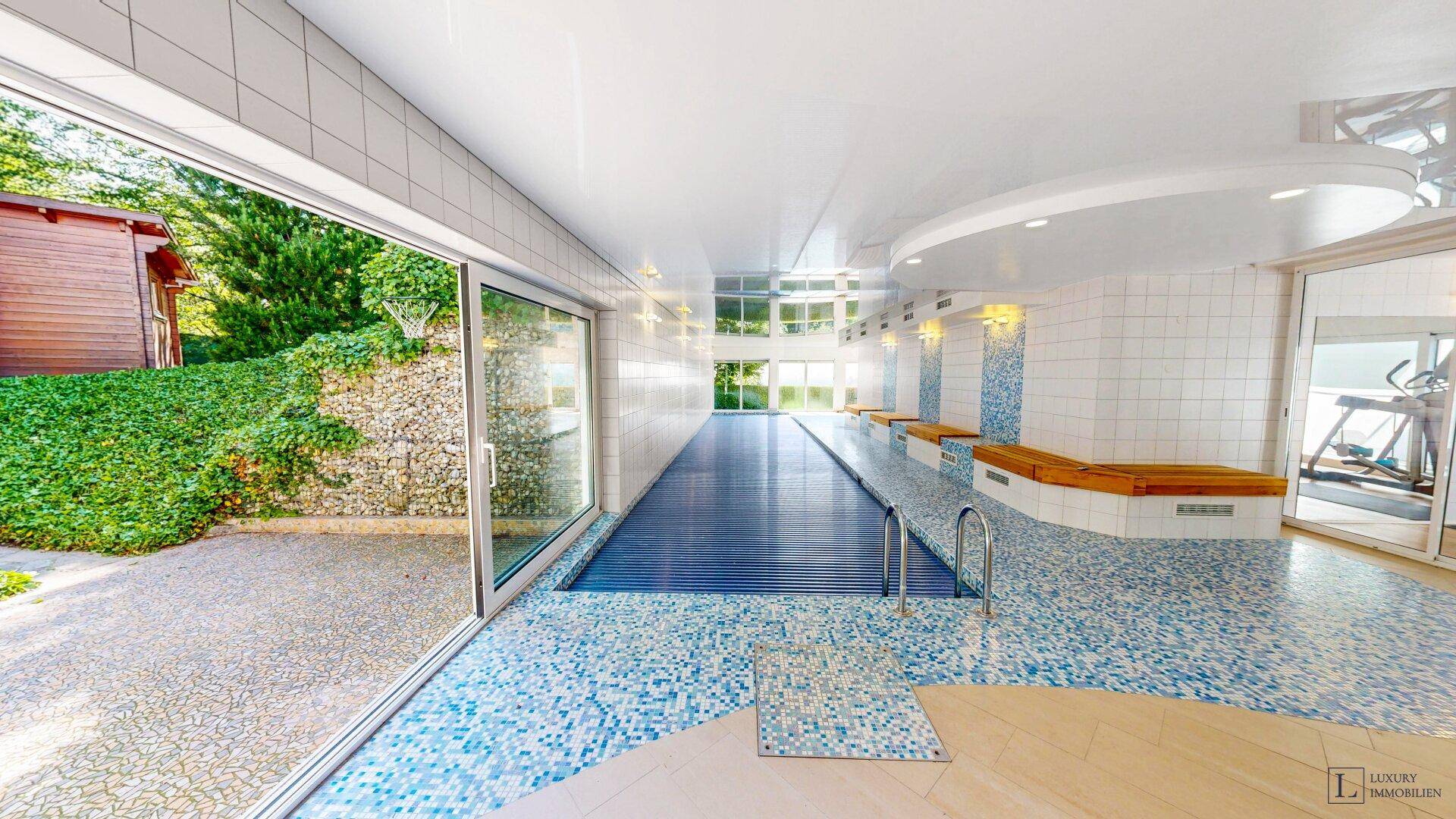 Großzügiger Innenpoolbereich mit blauen Mosaikfliesen und großen Fenstern zum Außenbereich.