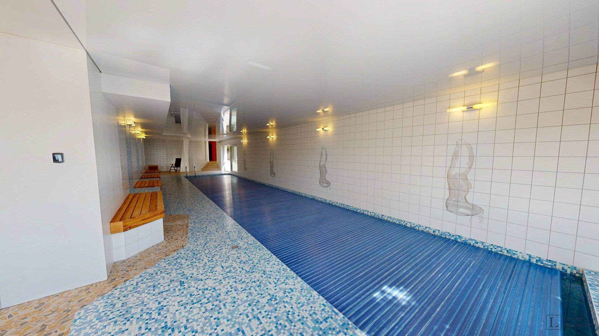 Langer Innenpool mit blauer Abdeckung, umgeben von weißen Fliesen und Holzbänken.