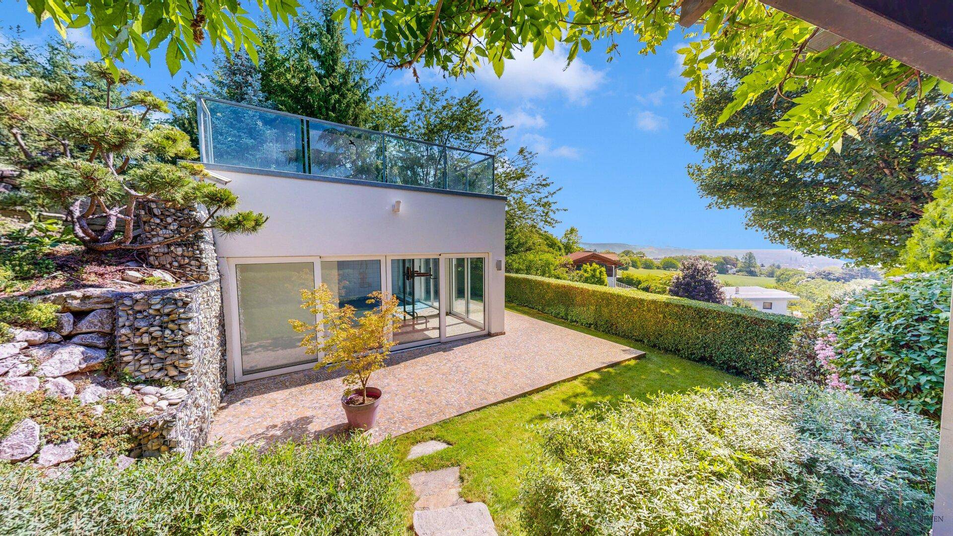 Untere Terrasse mit Kiesbelag und Zugang zum Garten, umgeben von üppiger Vegetation.