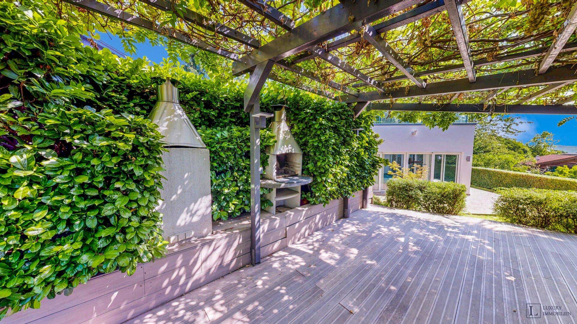 Gemütlicher Grillbereich auf einer Holzterrasse mit Pergola, umgeben von grünen Pflanzen.
