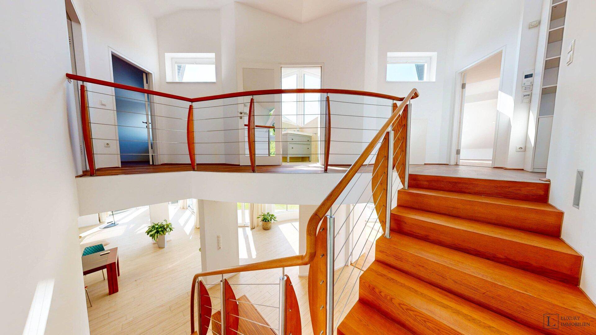 Elegante Holztreppe mit Edelstahlgeländer führt zu einem hellen Flur im Obergeschoss.