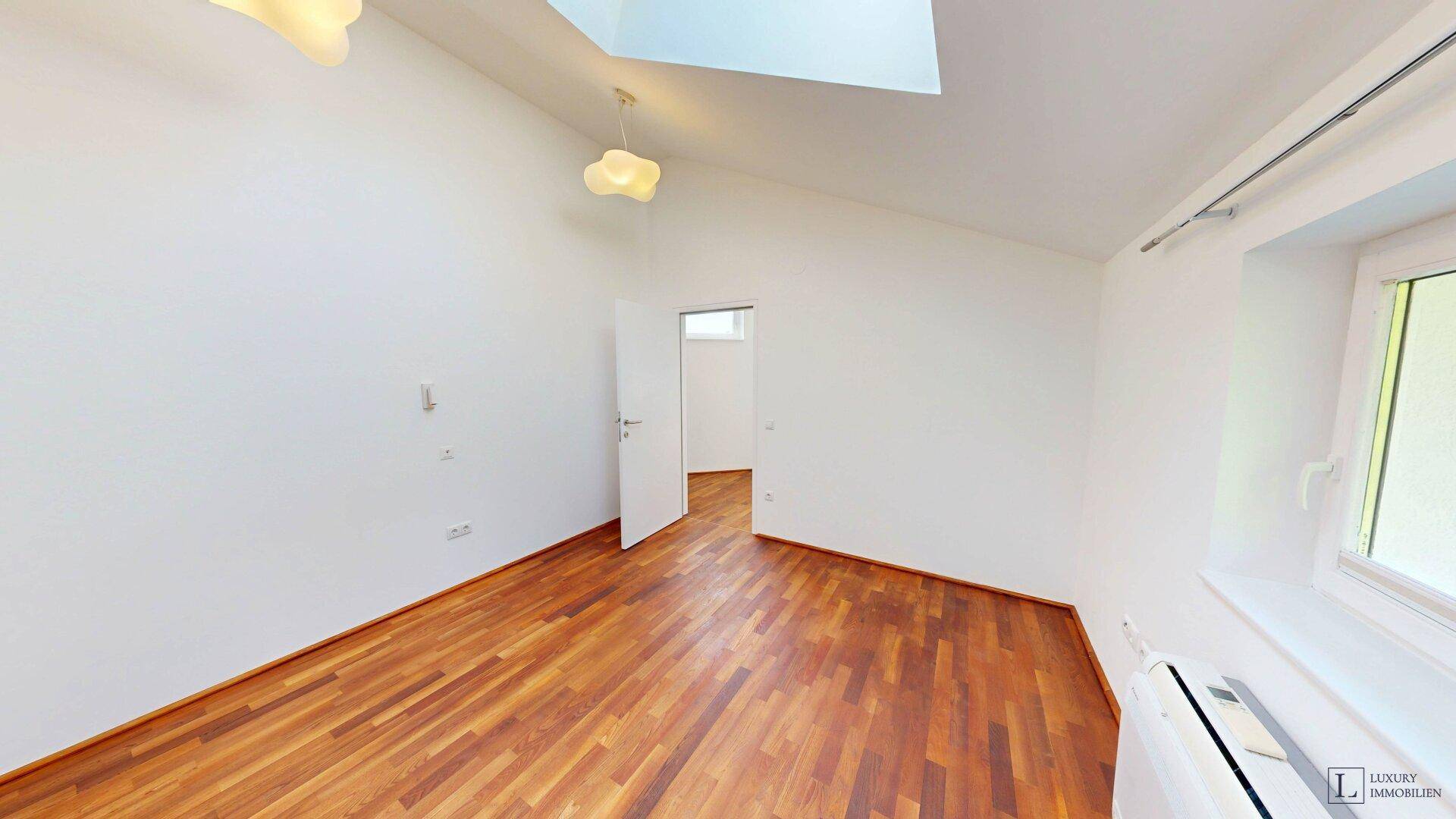 Leeres Zimmer mit Holzboden, Dachfenster und einem weiteren Fenster für viel Tageslicht.