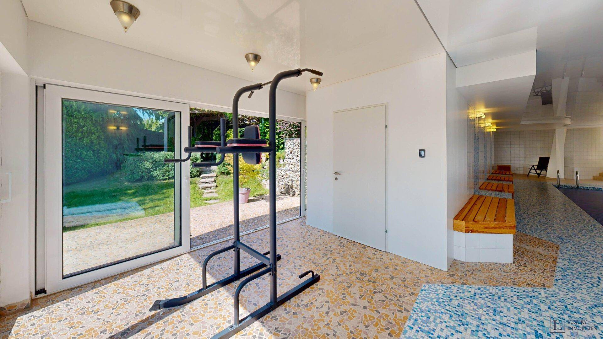 Heller Fitnessbereich mit Trainingsgerät, großen Fenstern und Blick in den grünen Garten.