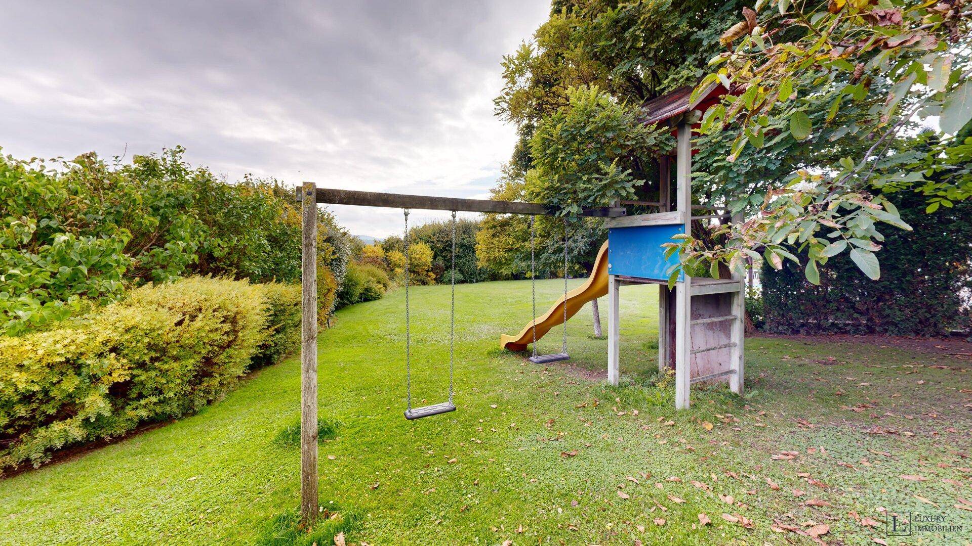 Kinderspielplatz im Garten mit Schaukel und Rutsche, umgeben von viel Grün.