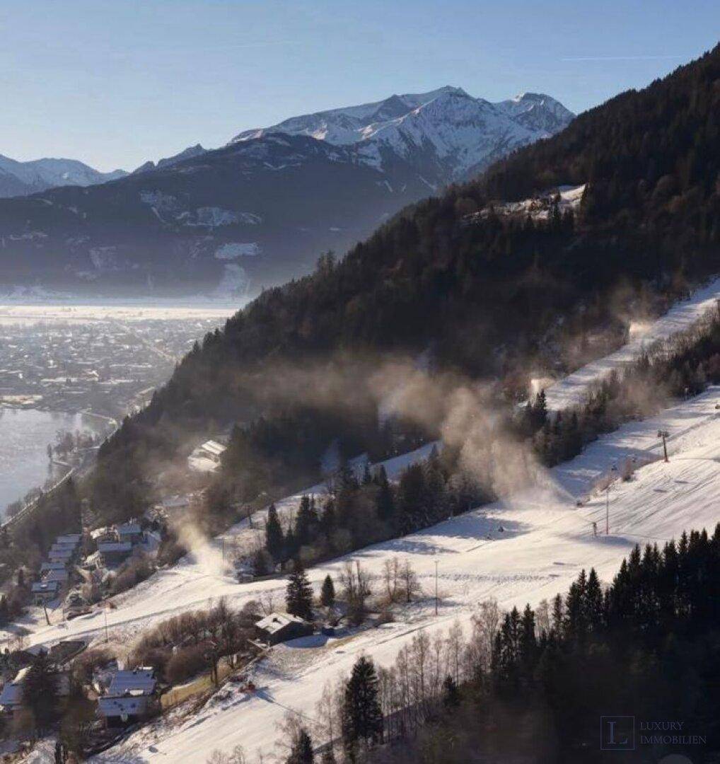 Winterliche Berglandschaft mit Skipiste, beschneiten Hängen und einem Tal mit See im Hintergrund.
