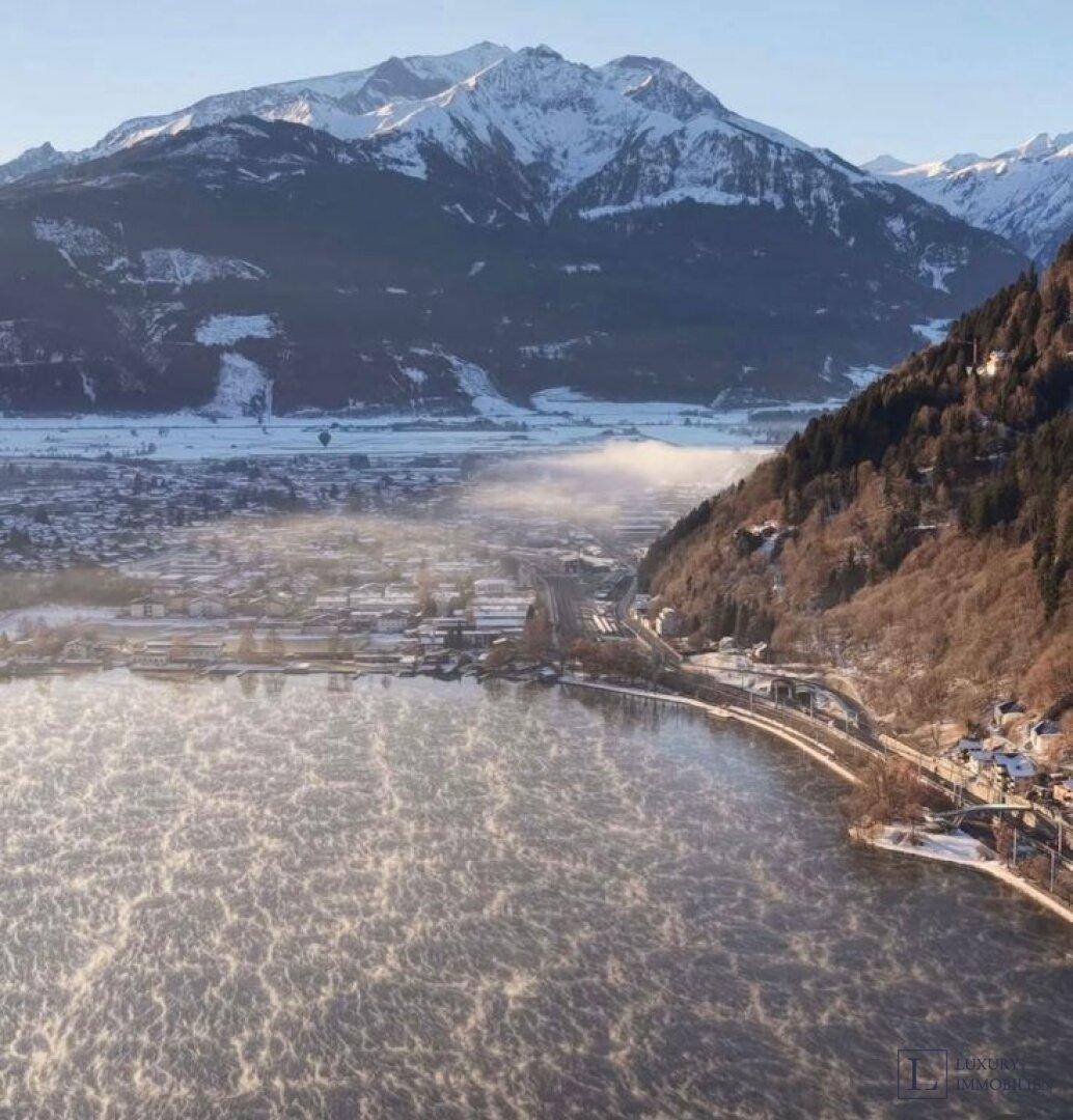 Panoramablick auf einen nebelverhangenen See, eine Stadt am Ufer und schneebedeckte Berge im Hintergrund.