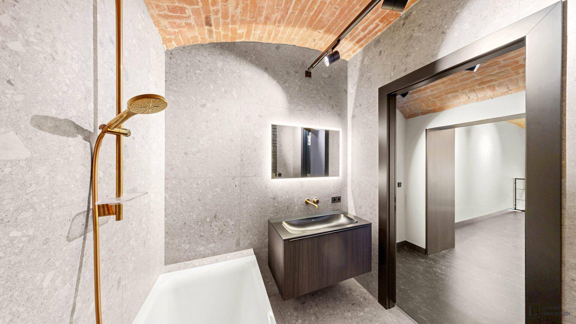 Modernes Badezimmer mit grauen Fliesen, freistehender Badewanne und goldfarbenen Armaturen.