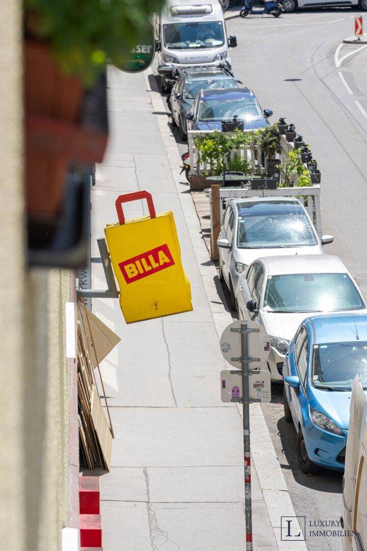 Belebtes Straßenbild mit parkenden Autos und einem großen gelben BILLA-Einkaufstaschen-Werbeobjekt.