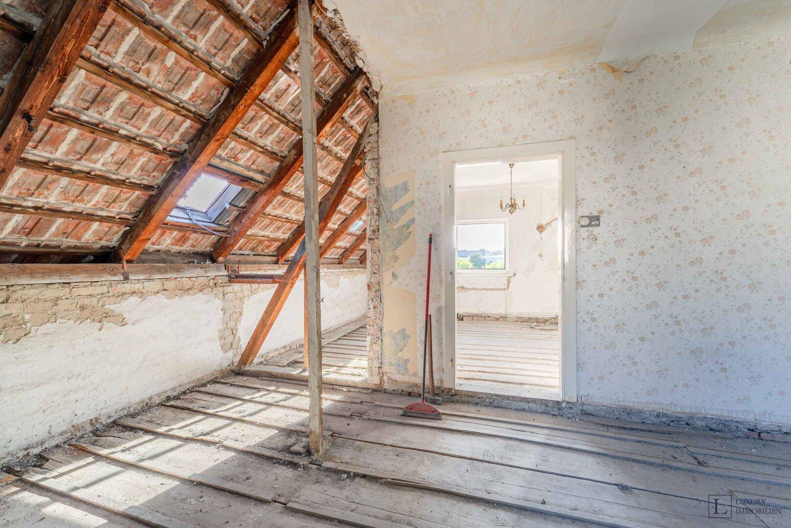 Dachbodenraum im Rohzustand mit sichtbaren Dachbalken und einem Dachfenster für natürliches Licht.