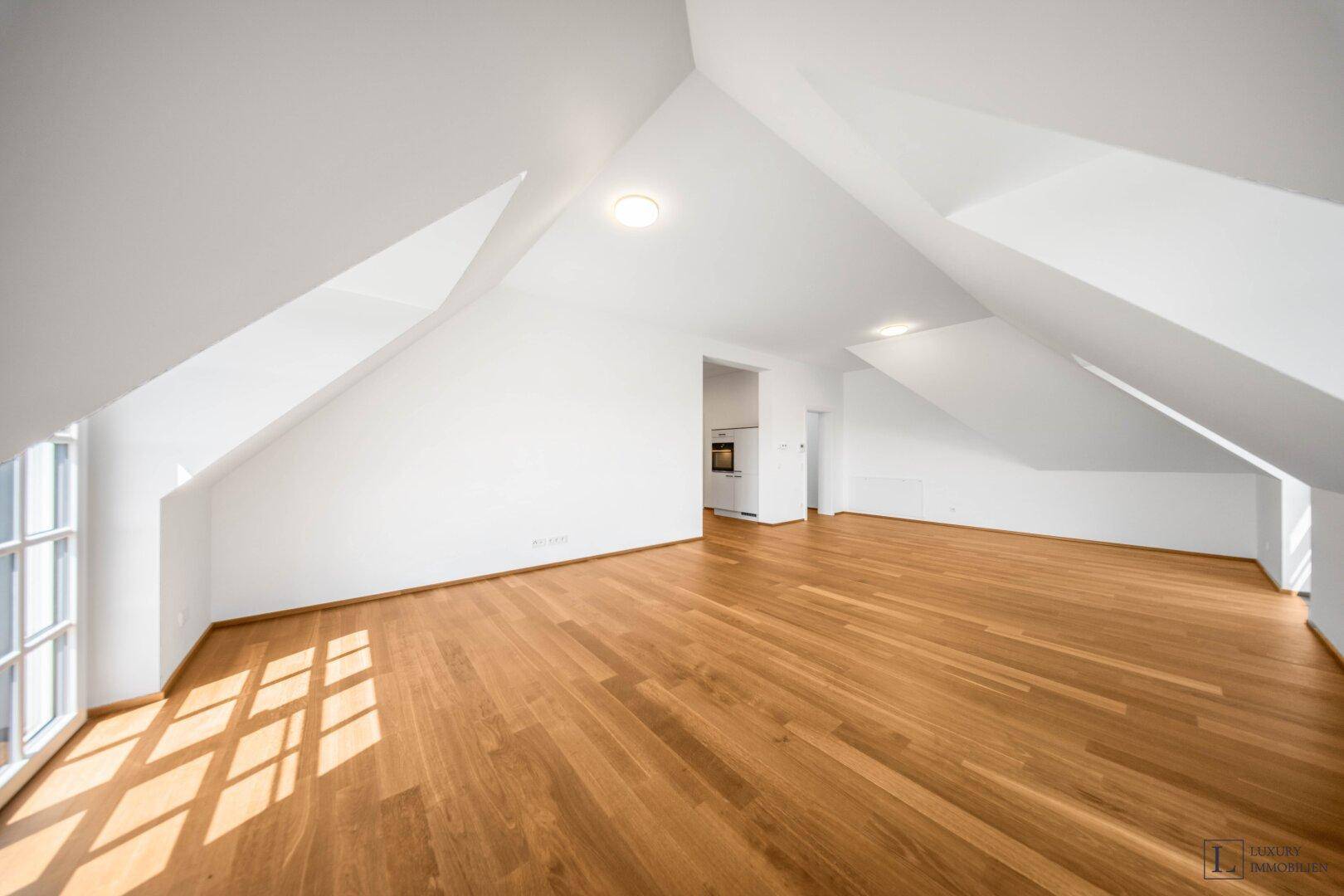 Großer, heller Raum mit Holzboden, Dachschrägen und Blick auf den Küchenbereich.