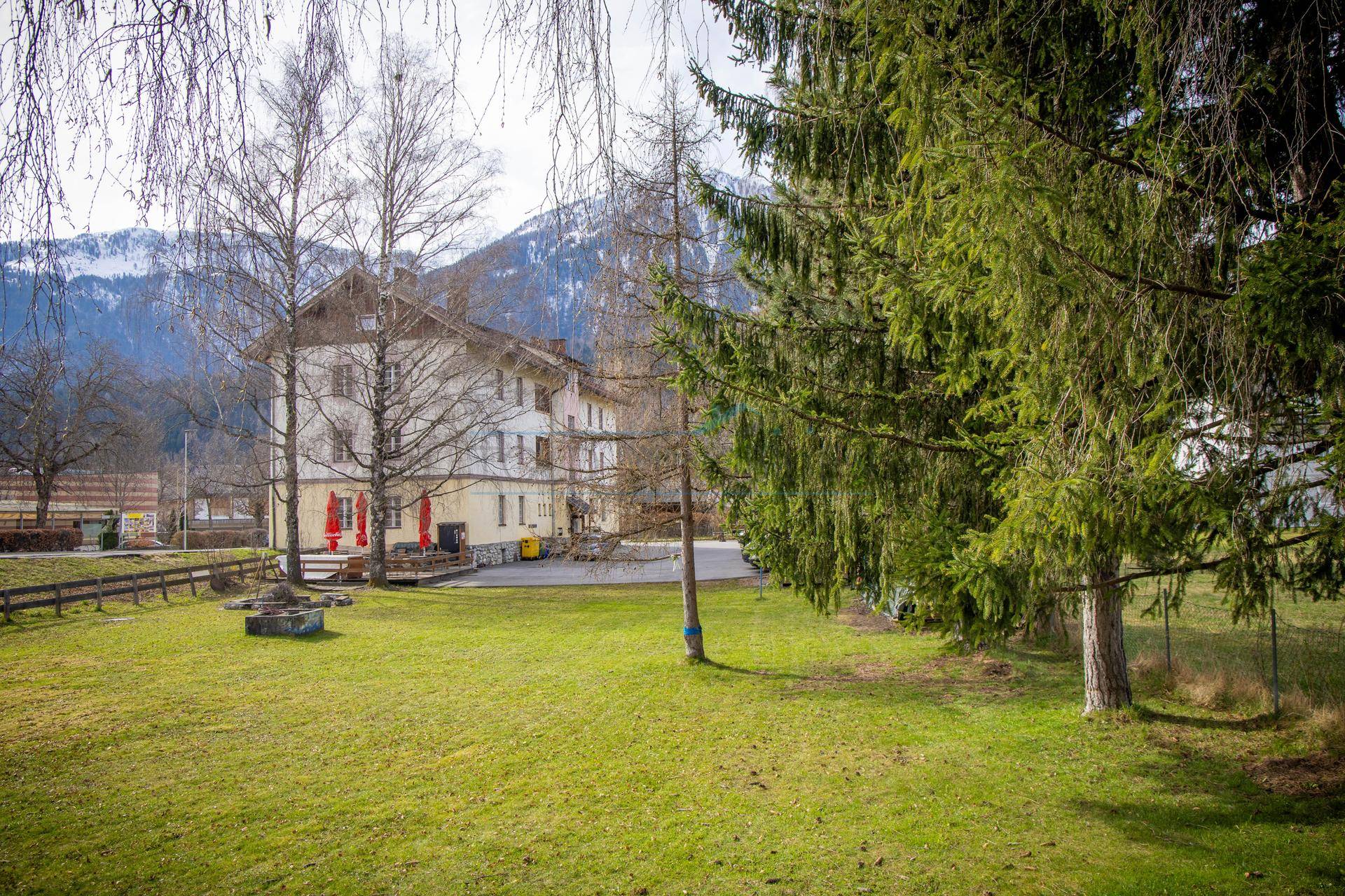 Ansicht des Gebäudes von einer grünen Wiese aus, mit Bäumen im Vordergrund und Bergen im Hintergrund.