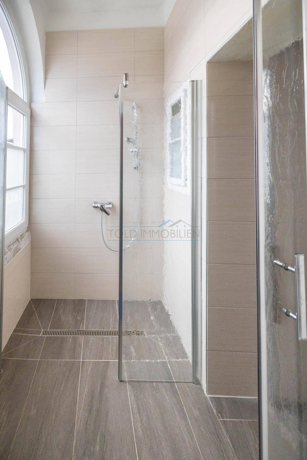 Modernes Badezimmer mit begehbarer Dusche, hellen Wandfliesen und dunkleren Bodenfliesen, sehr funktional.
