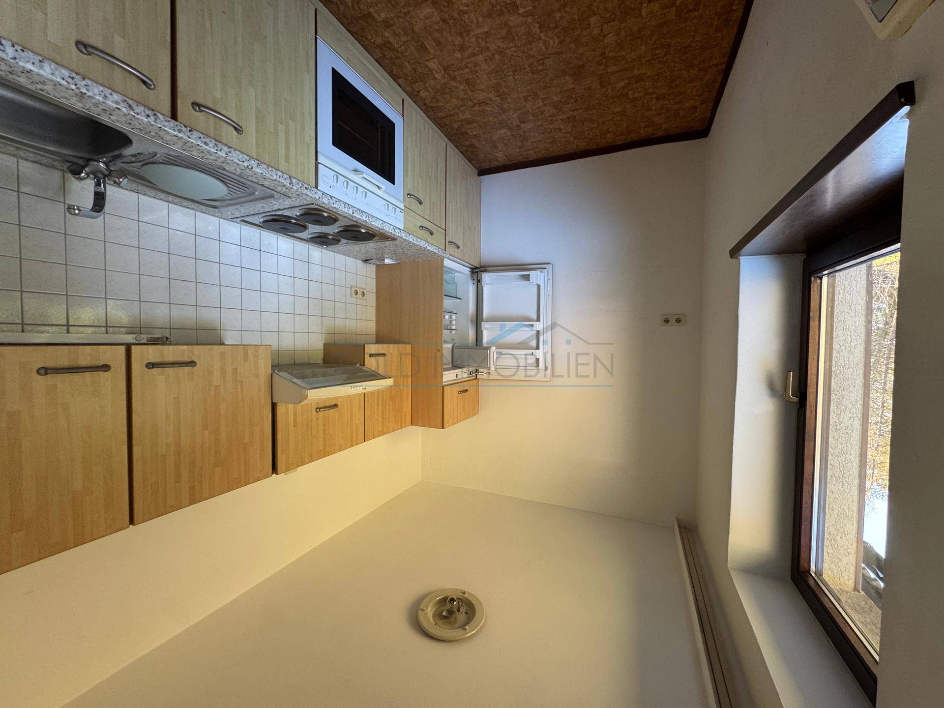 Kleine Küche mit Holzschränken, Elektroherd und Fenster, das Tageslicht hereinlässt.