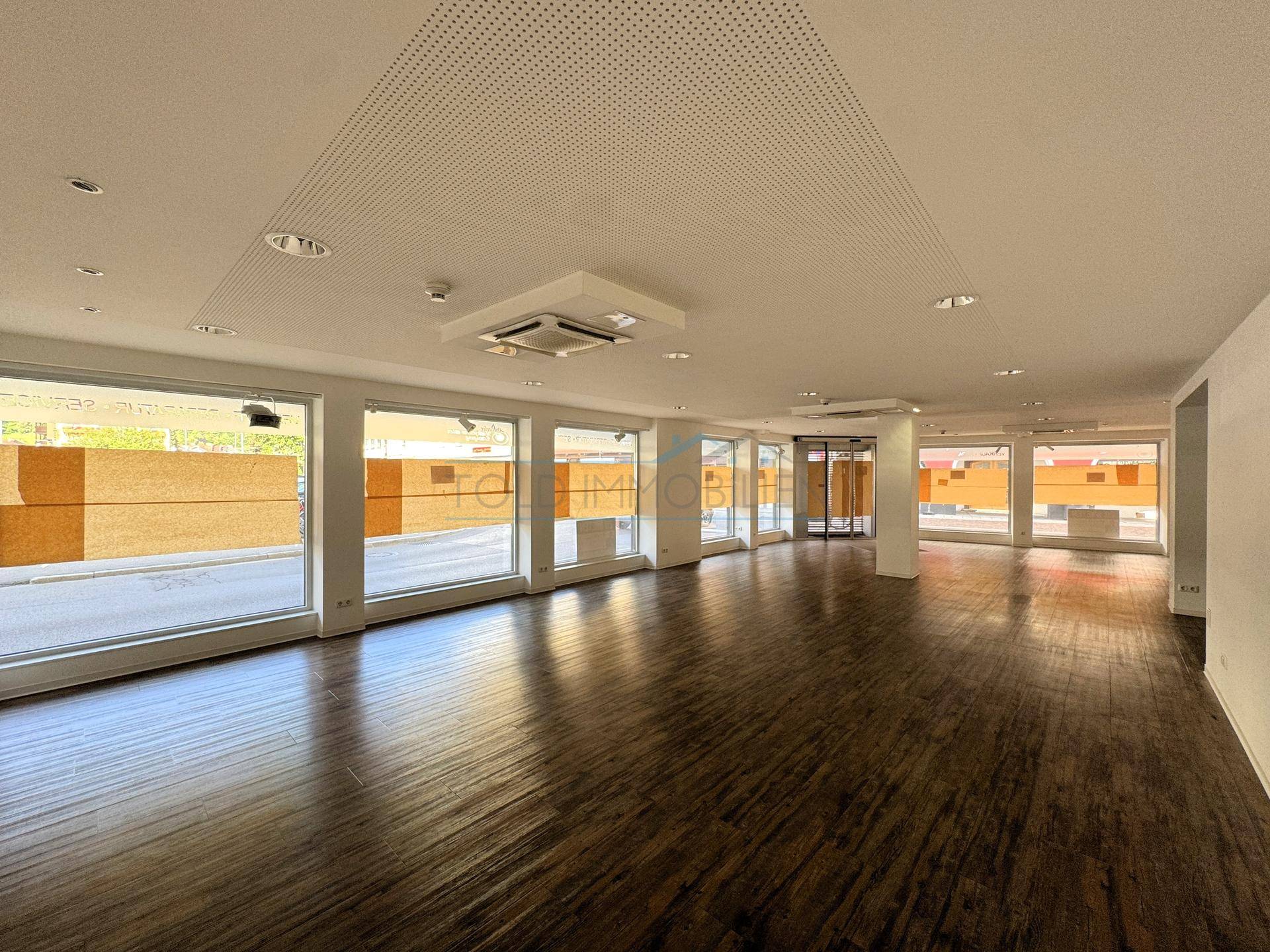 Geräumiger, leerer Ladenbereich mit dunklem Holzfußboden, hohen Decken und großen Schaufenstern für viel Tageslicht.